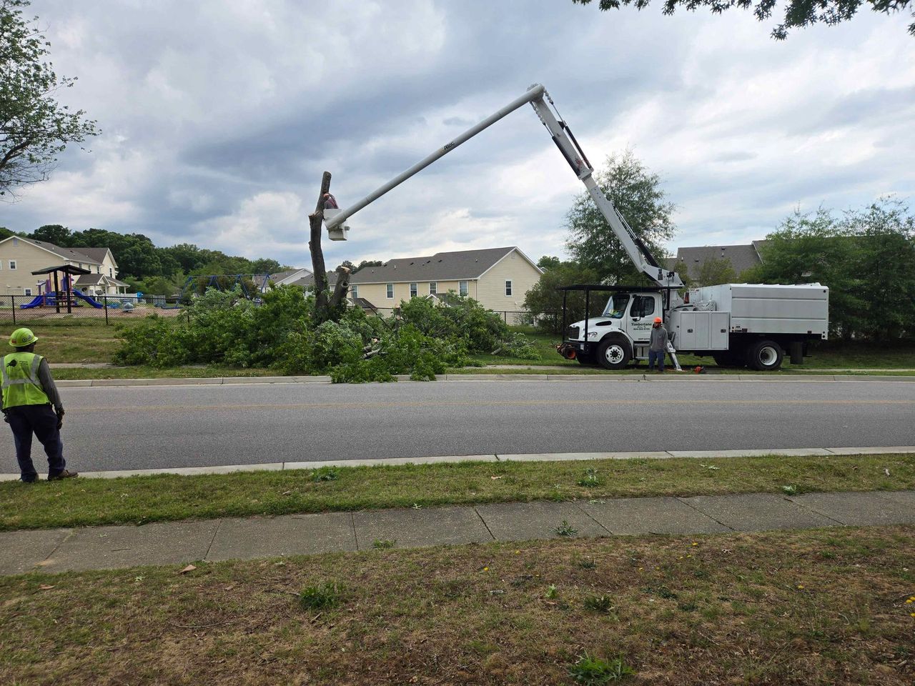 Utility trucks trimming storm-damaged trees with a bucket crane on a roadside in a suburban neighborhood Utility trucks trimming storm-damaged trees with a bucket crane on a roadside in a suburban neighborhood