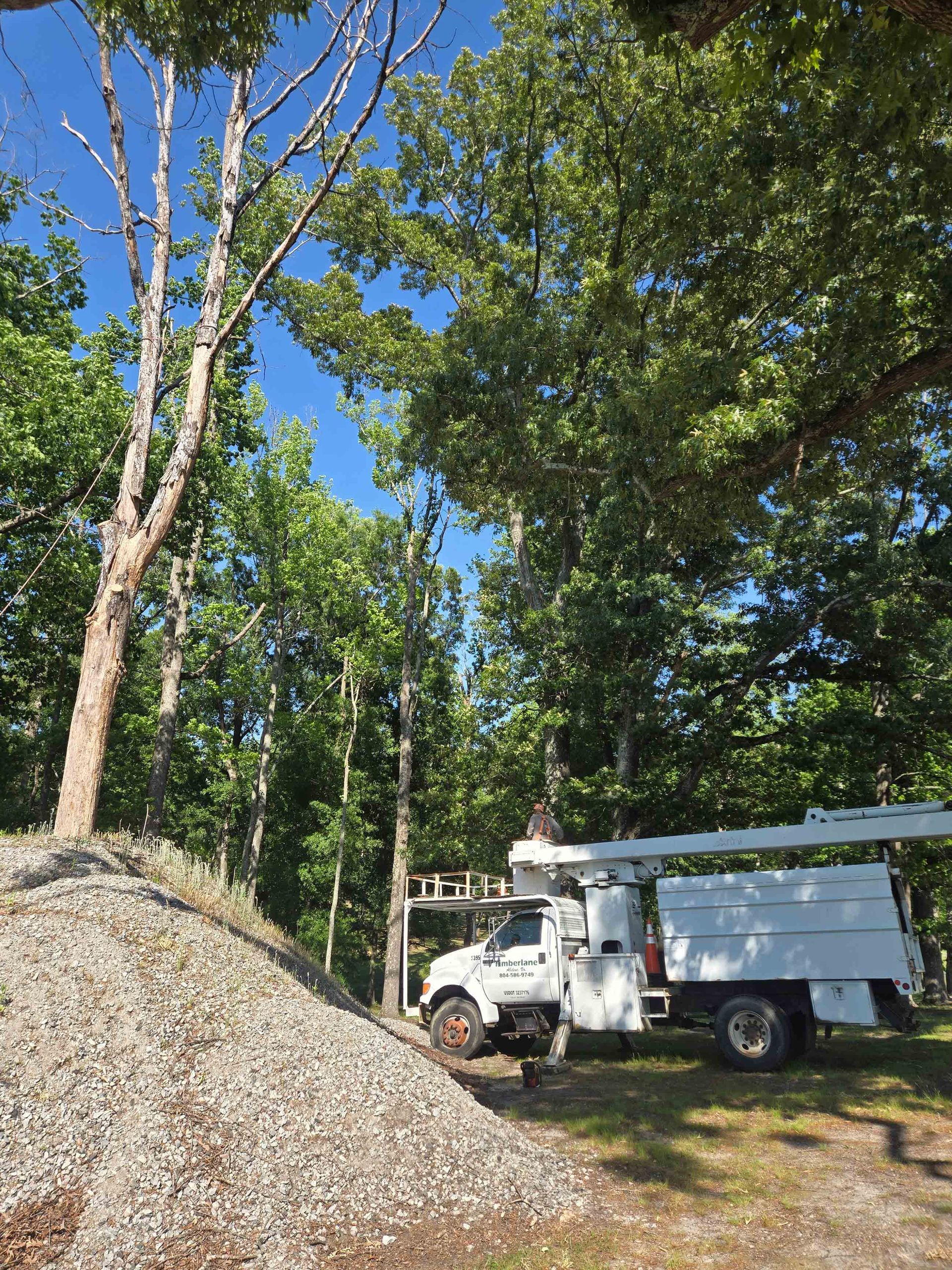 White utility truck parked beside gravel pile in a wooded area under a blue sky