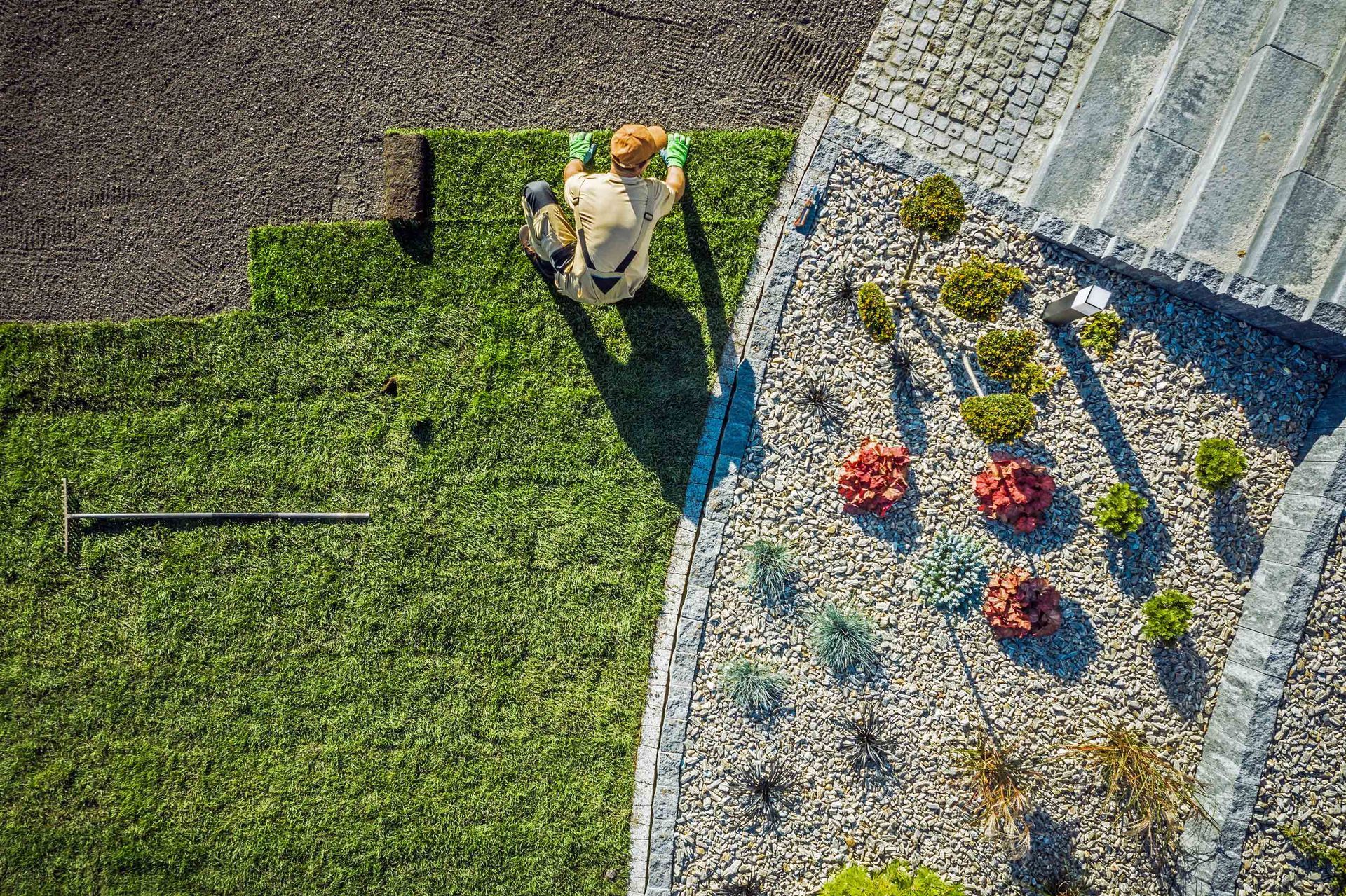 Person in a yellow hard hat works near a white building, beside green lawn and flower beds.