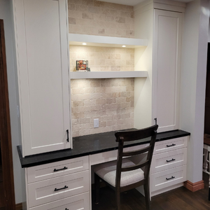 A white home office built-in featuring a black desk, drawers, and illuminated shelving set against a brick backsplash.