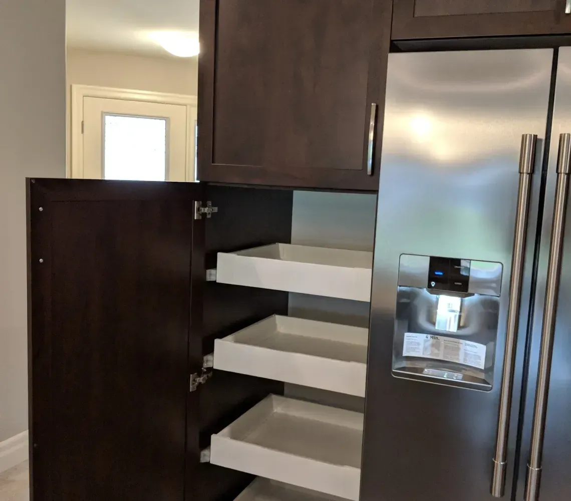 A dark wood kitchen cabinet with the door open, revealing four white slide-out pantry shelves next to a stainless fridge.