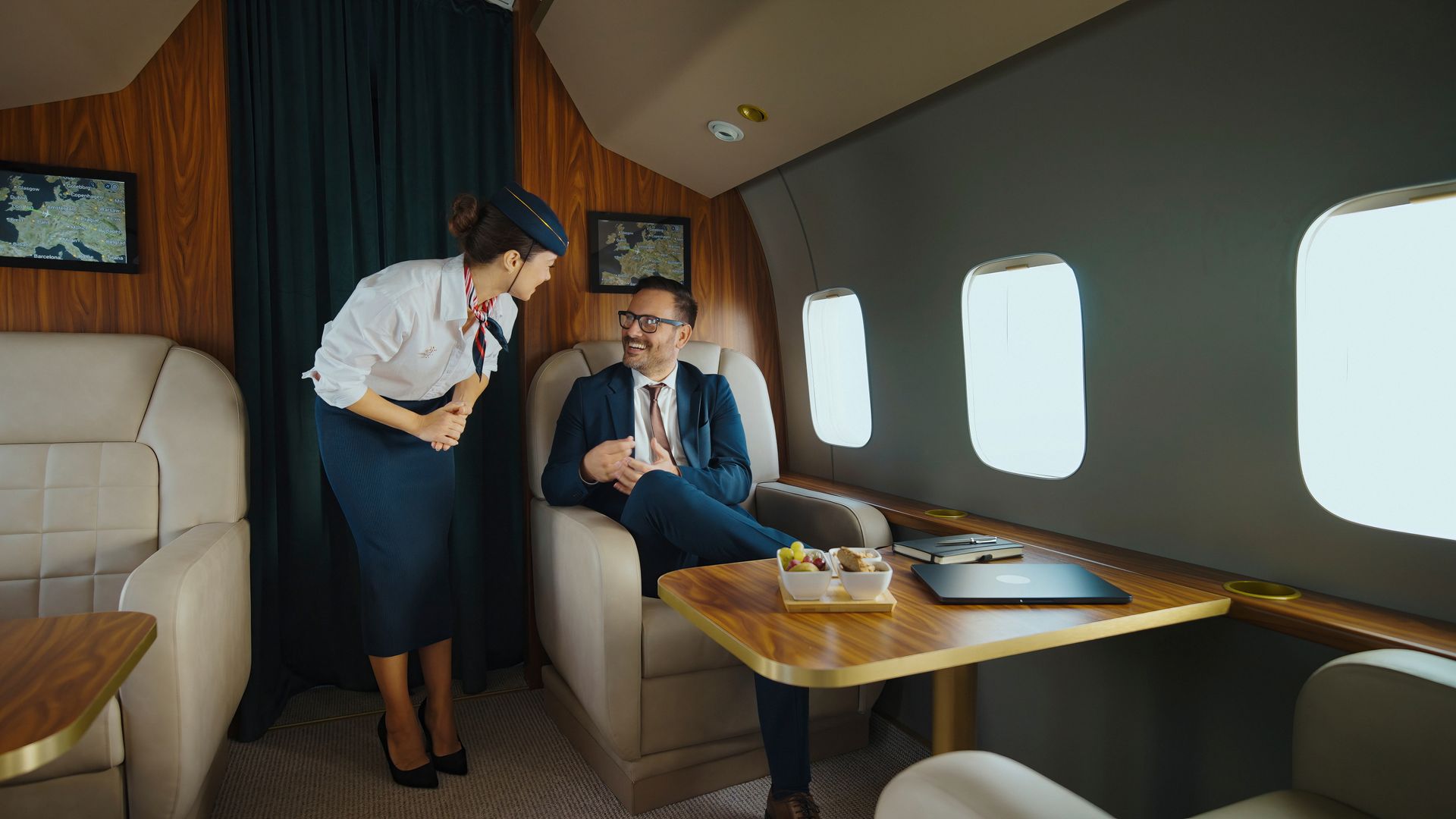 Two people seated in a private jet cabin, one standing and serving drinks beside a small table.