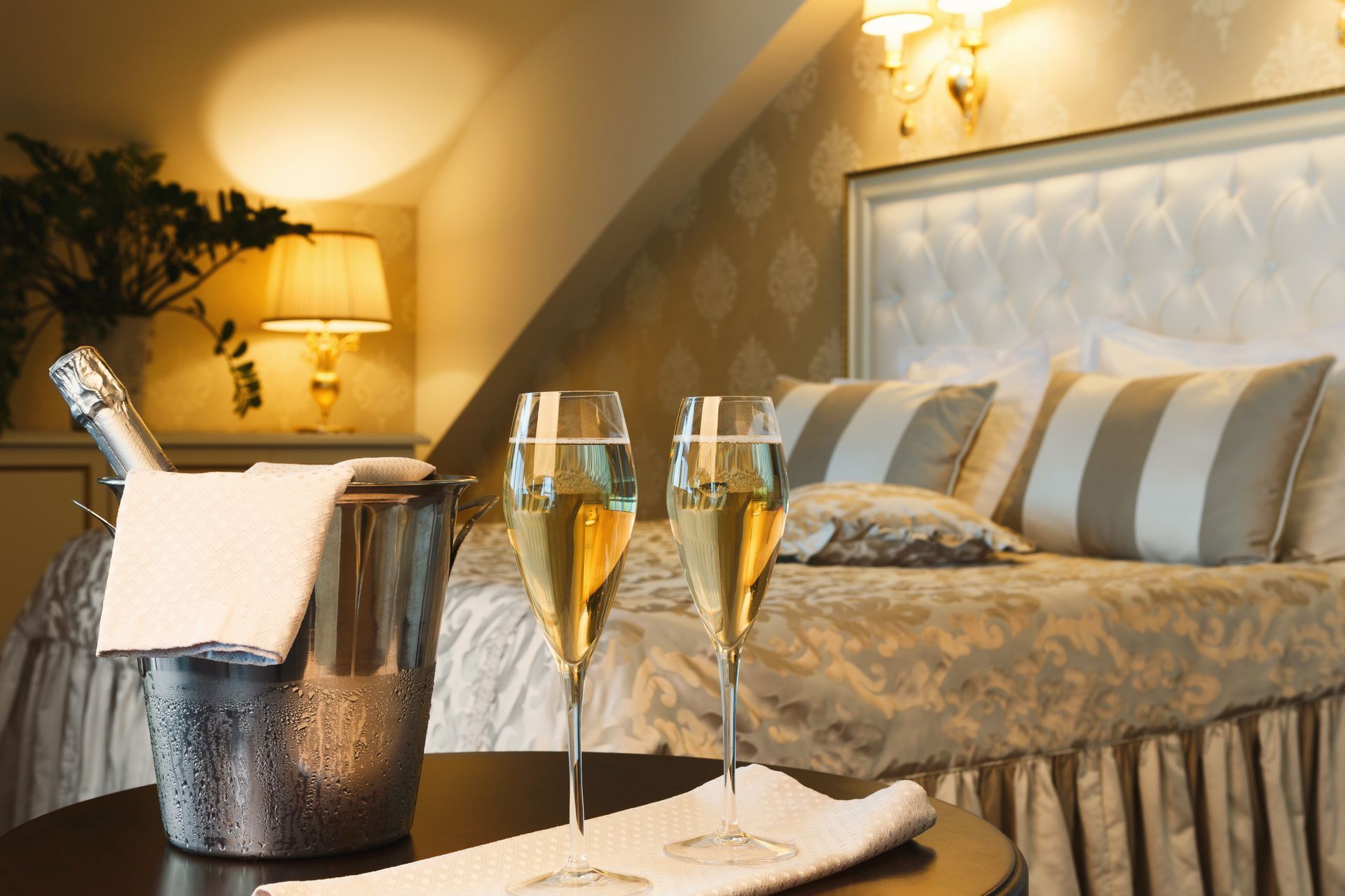 Romantic hotel bed with champagne on ice and two filled flutes on a tray, warm lamp light