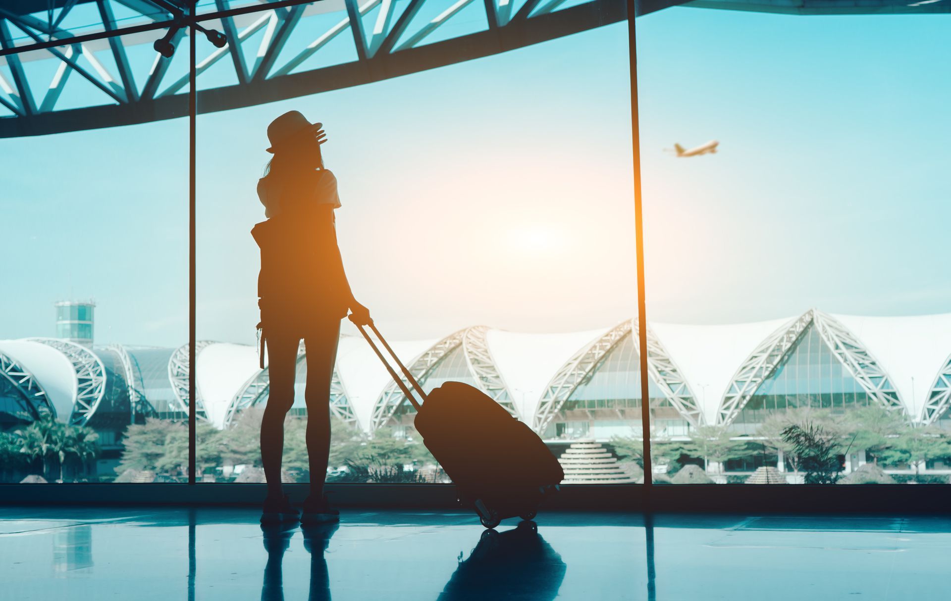 Silhouette with rolling suitcase at airport window, planes and terminals in sunrise light