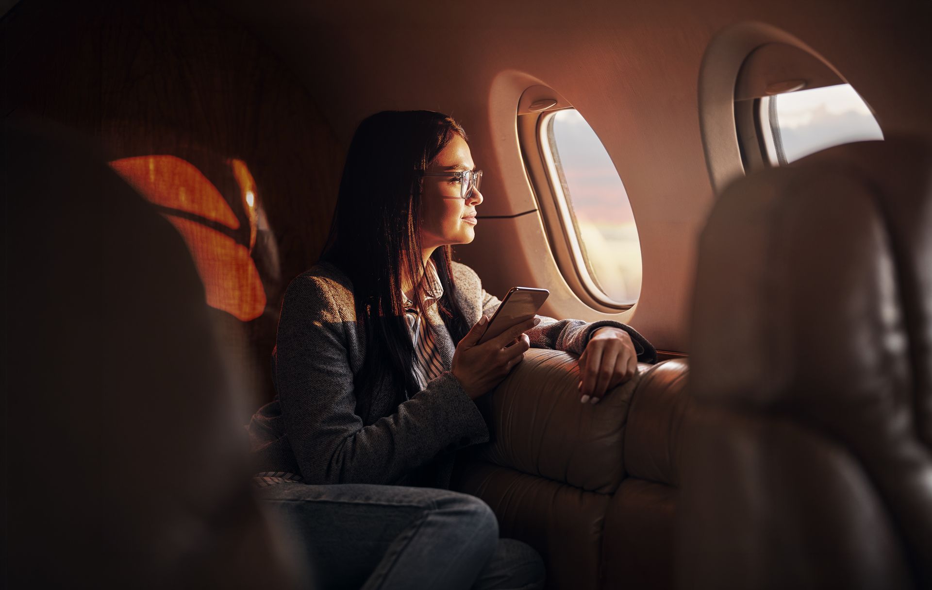 Passenger seated by an airplane window, holding a phone and looking outside at sunset.