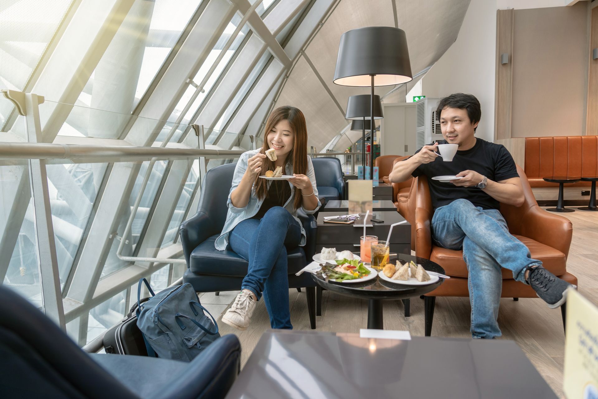 Two people sitting in a modern lounge, eating and using phones beside a small table with food and drinks.