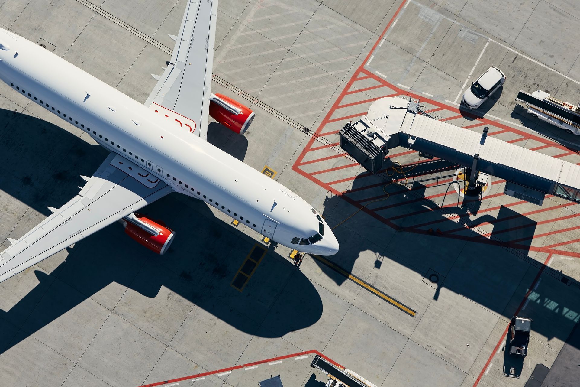 Top-down view of a passenger jet at an airport gate beside a jet bridge and service vehicles.