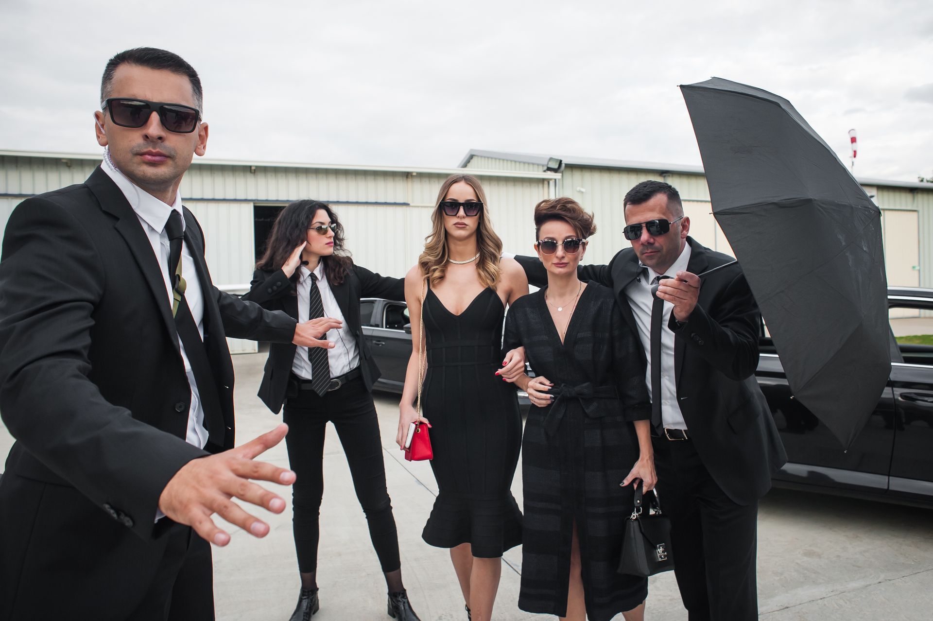 Five people in black formalwear posing beside a luxury car with an open door, one holding an umbrella
