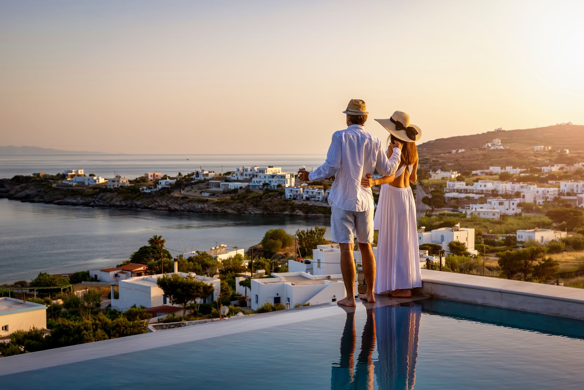 Two people standing by an infinity pool overlooking a coastal town at sunset