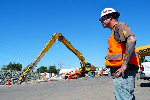 A construction worker stands in front of a large yellow crane that says nor