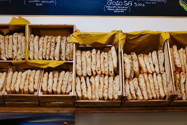 Esposizione di baguette in vassoi di legno, alcune avvolte in carta gialla, con una lavagna sopra.