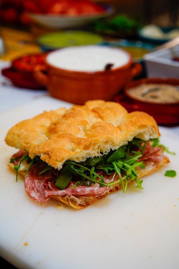 Panino al forno con verdure e affettati su un piatto bianco, con piatti colorati sfocati sullo sfondo.
