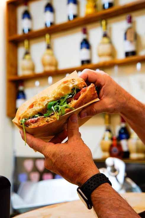 Mani che reggono un panino in un bar, con scaffali di bottiglie sfocati sullo sfondo.