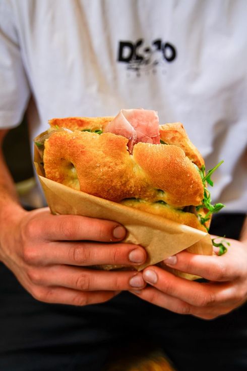 Mani che reggono un toast con ripieno rosa e verdure, indossano una camicia bianca e pantaloni neri.