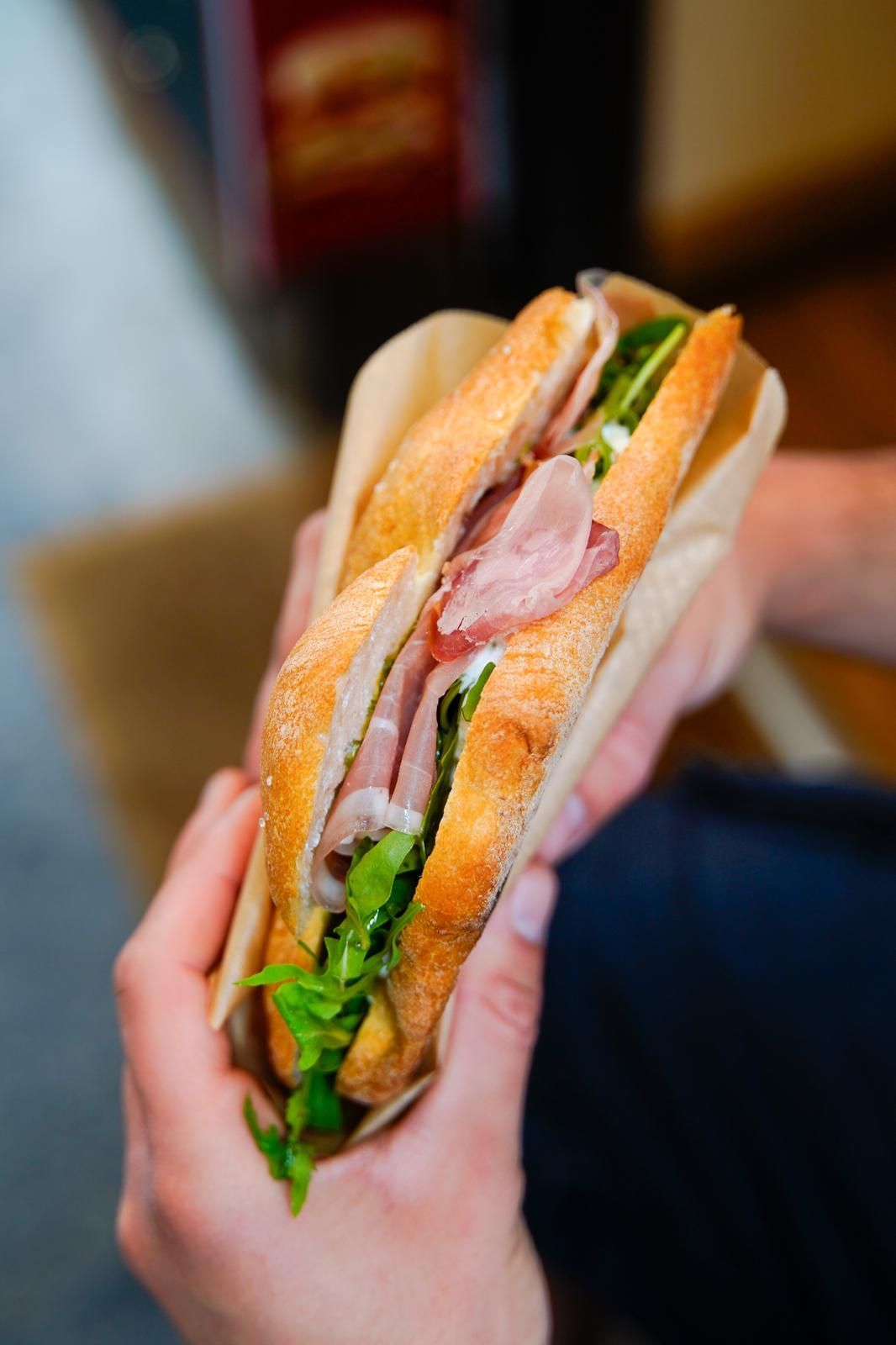 Mani che reggono un panino con lattuga, pomodoro, prosciutto e pane bianco.