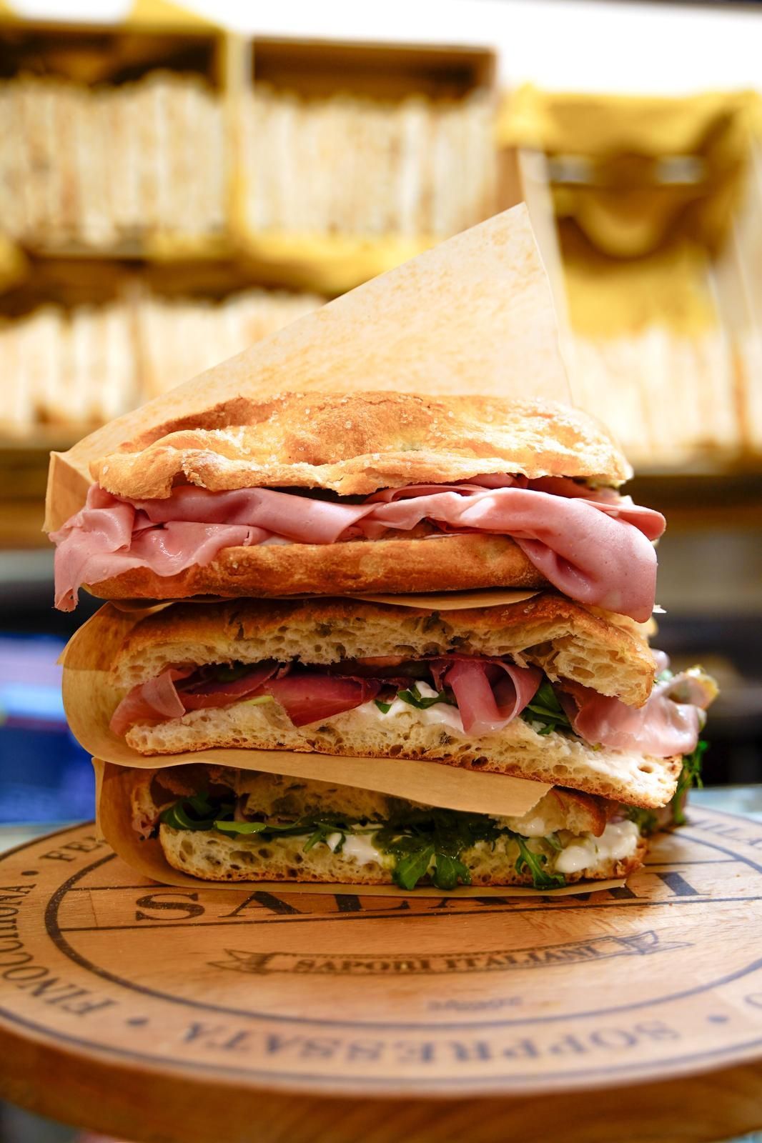 Pila alta di sandwich con affettati e verdure su un tagliere di legno, su uno sfondo sfocato.