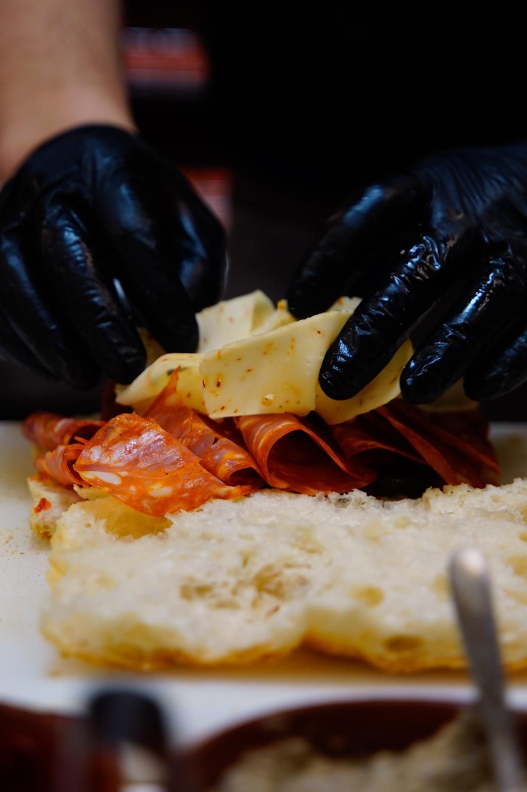 Mani guantate che assemblano un panino con carne e formaggio piegati su una fetta di pane.