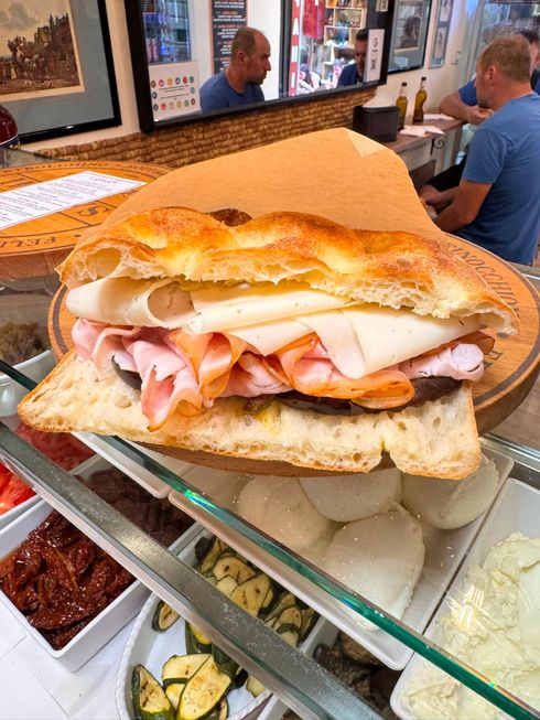 Mani che reggono un panino con lattuga, pomodoro, prosciutto e pane bianco.