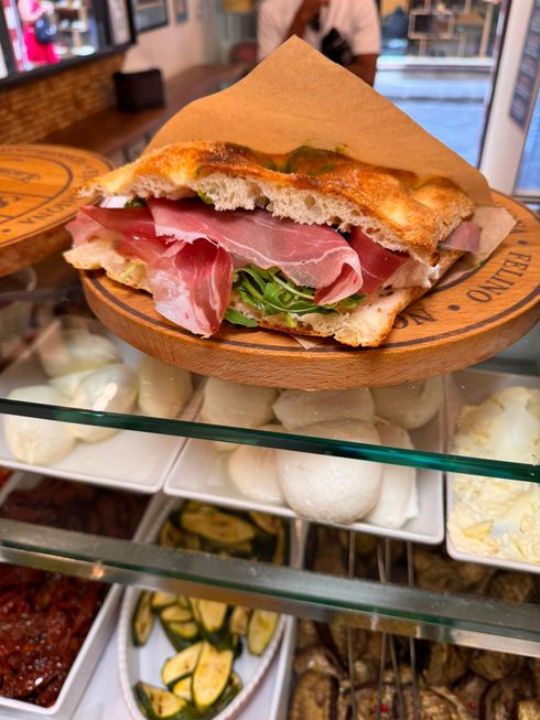 Mani che reggono un panino con lattuga, pomodoro, prosciutto e pane bianco.