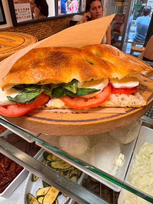 Mani che reggono un toast con ripieno rosa e verdure, indossano una camicia bianca e pantaloni neri.