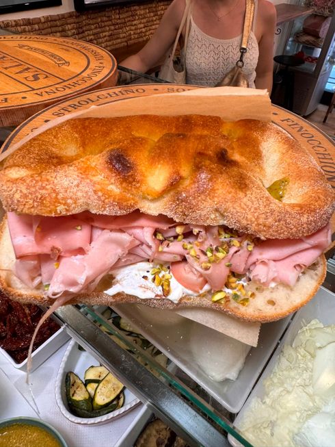 Mani che reggono un panino con lattuga, pomodoro, prosciutto e pane bianco.