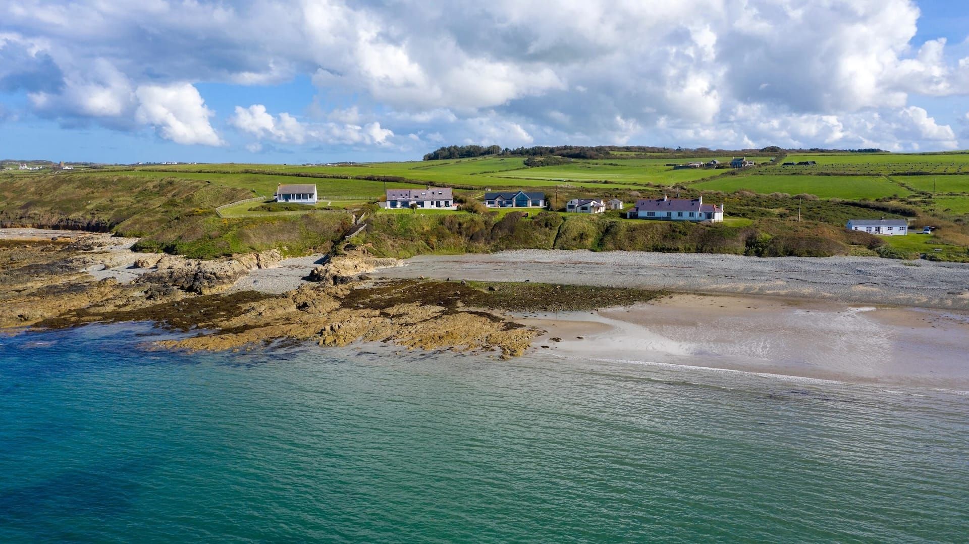 Coastal shoreline with turquoise water, rocky beach, and small houses beneath a green hillside under cloudy sky