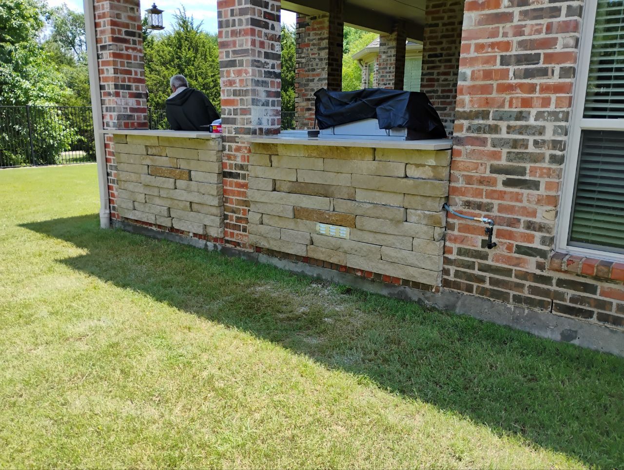 Stone and brick outdoor patio with a grill under a covered area and green grass.