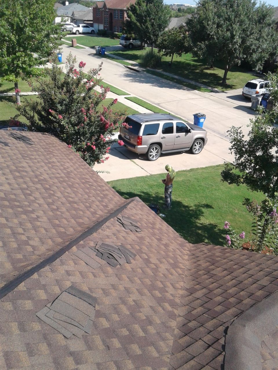 View of a brown roof with missing shingles, overlooking a suburban street with a parked SUV and green lawns.