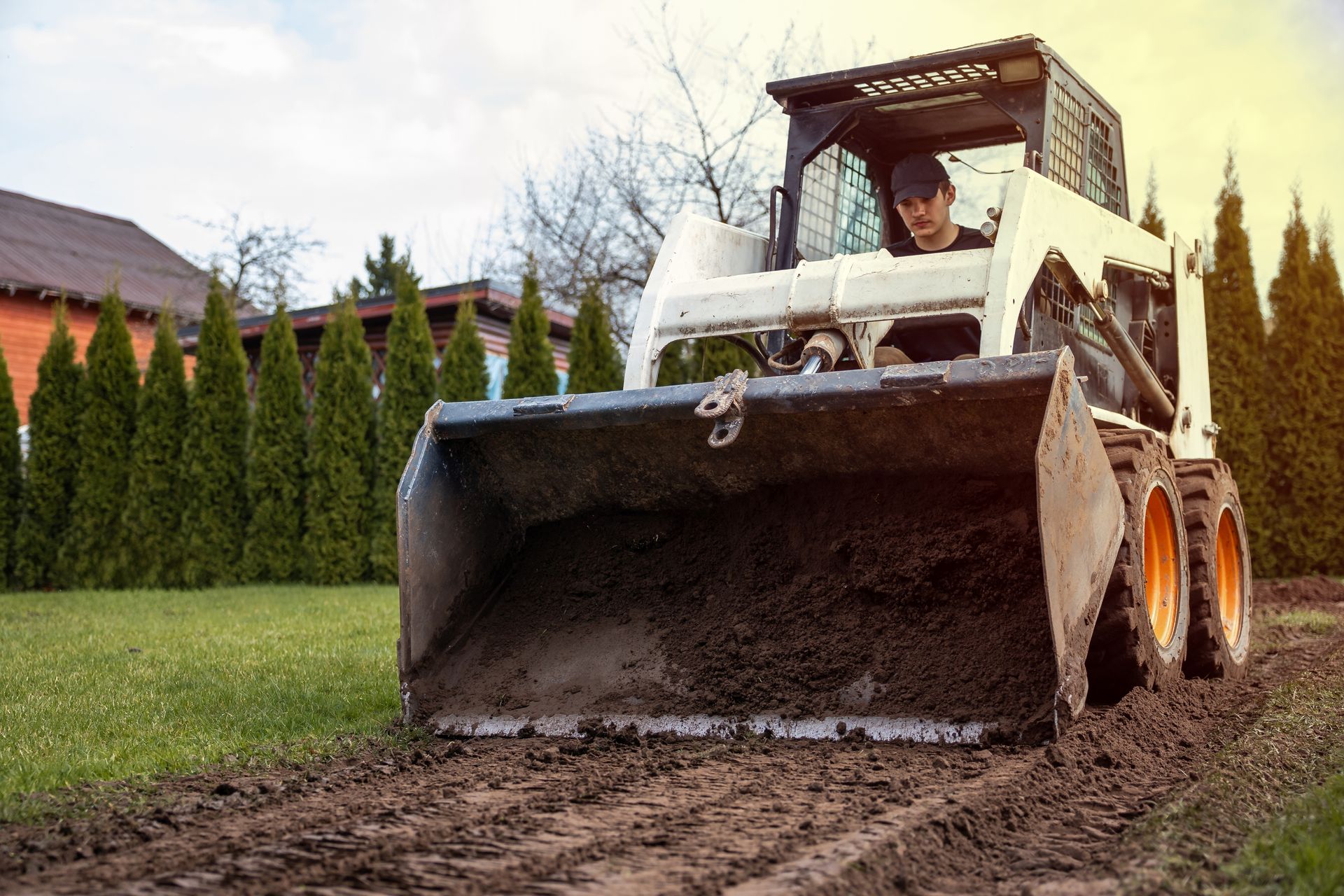 Front loader scooping mud on a grassy property beside hedges and a house