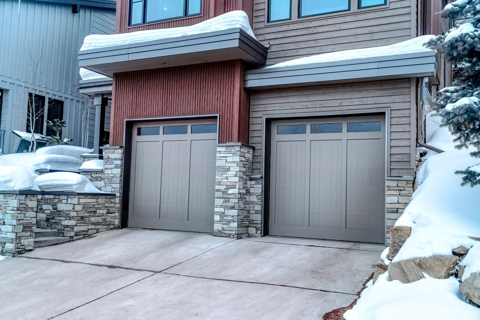 Snow-covered driveway and two garage doors beneath a house with stone and wood siding.