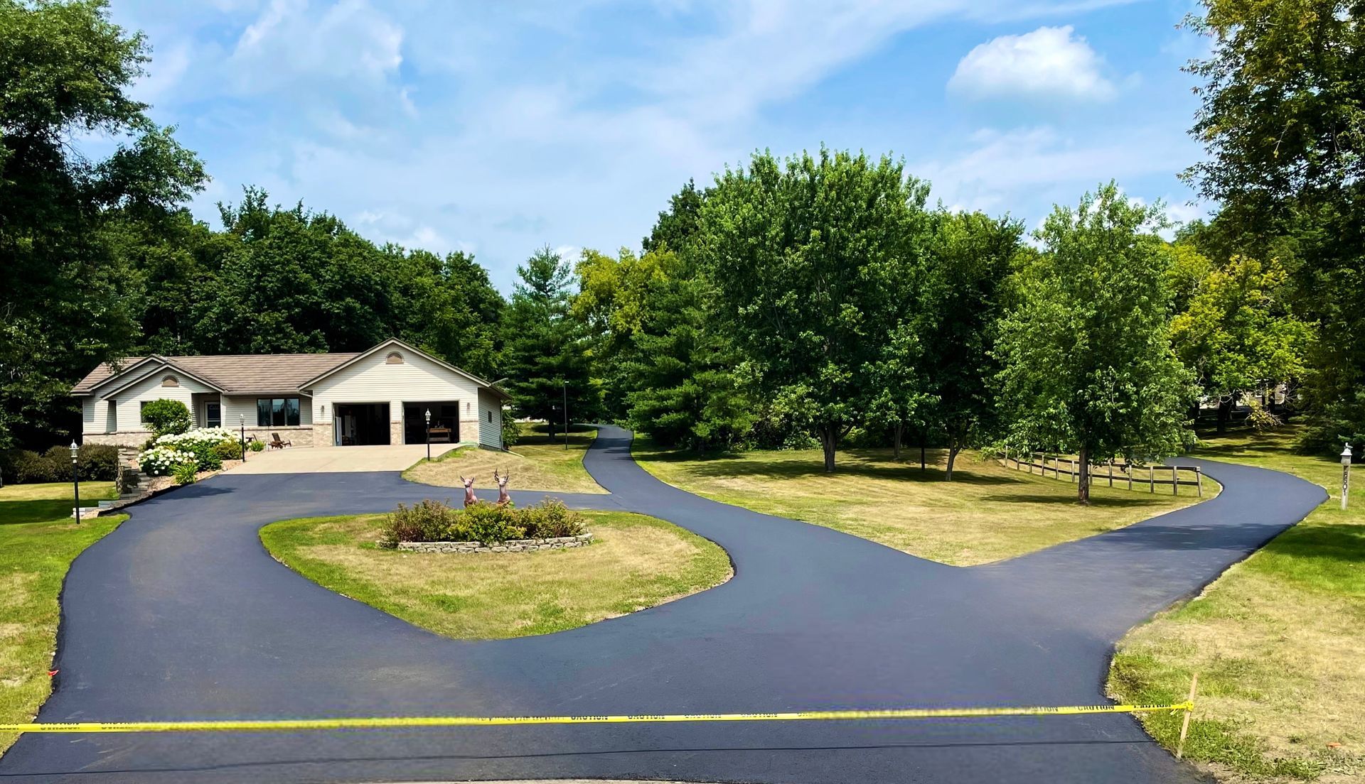 Driveway splits to reach a house, surrounded by trees and grass, on a sunny day.