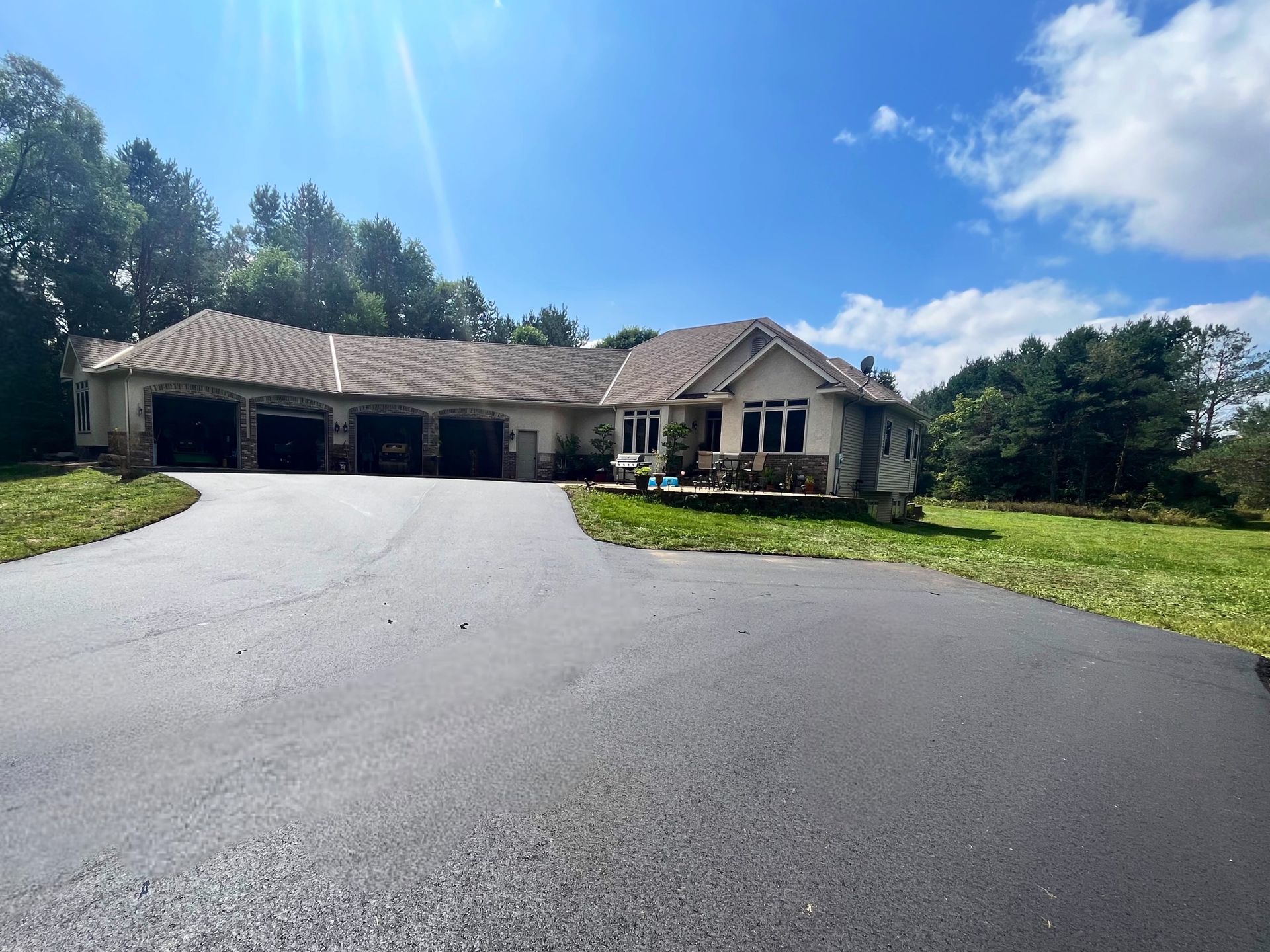 House with three-car garage, asphalt driveway, and trees under a blue sky with sun glare.