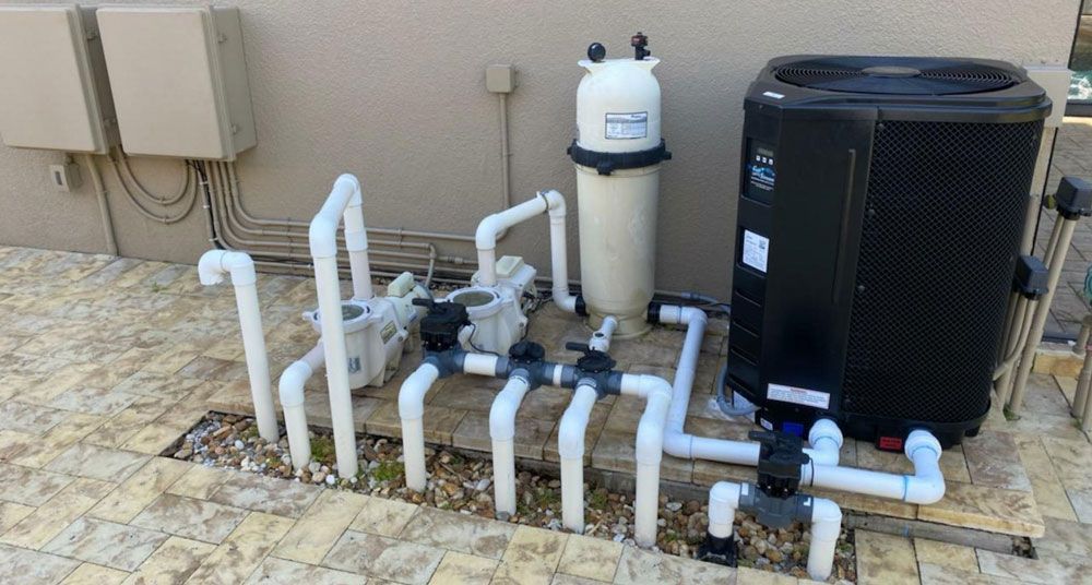 Pool equipment, including a pump, filter, and heat pump, connected by white PVC pipes, set on a tiled patio.