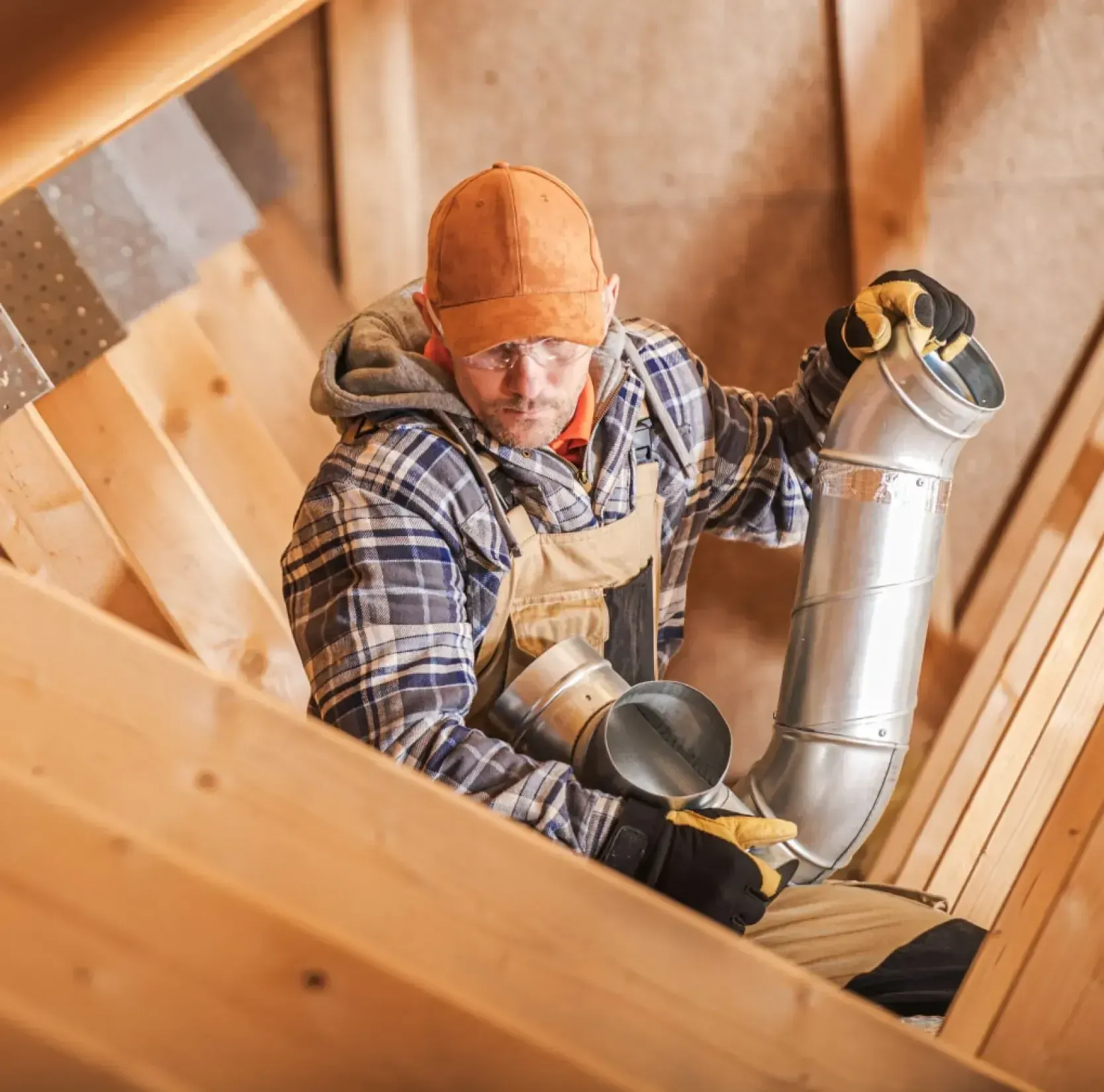 A worker in an attic installing ductwork, wearing gloves and a cap.