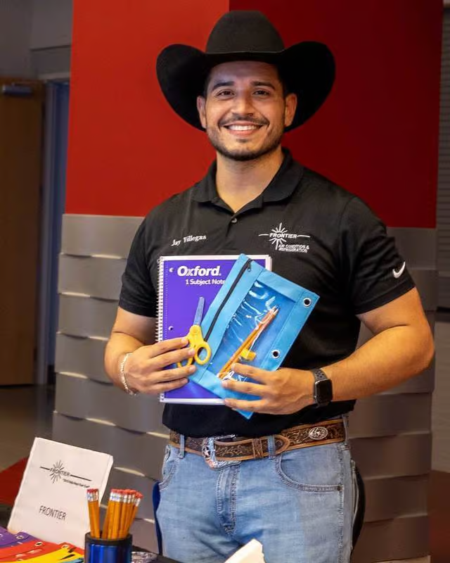 Man in cowboy hat holding school supplies, smiling. Black shirt, jeans, belt. Red wall backdrop.