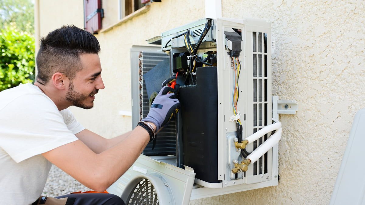 Man in gloves repairs an outdoor air conditioning unit on a beige building wall.