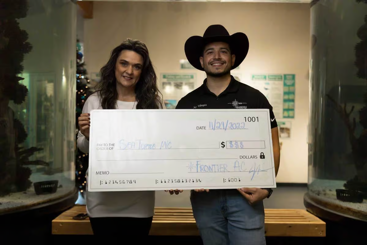 Two people holding a large check. Woman in white shirt, man in cowboy hat; fish tank in background.