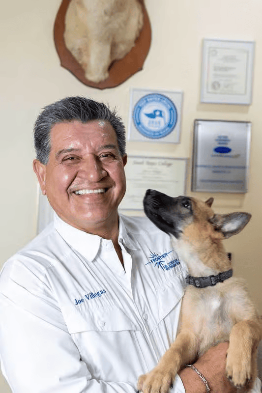 Man holding a puppy, smiling. White shirt, various plaques on the wall.