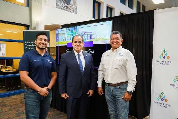 Three men stand in front of a screen displaying a building. Two men wear company shirts, one in a suit. Indoors.