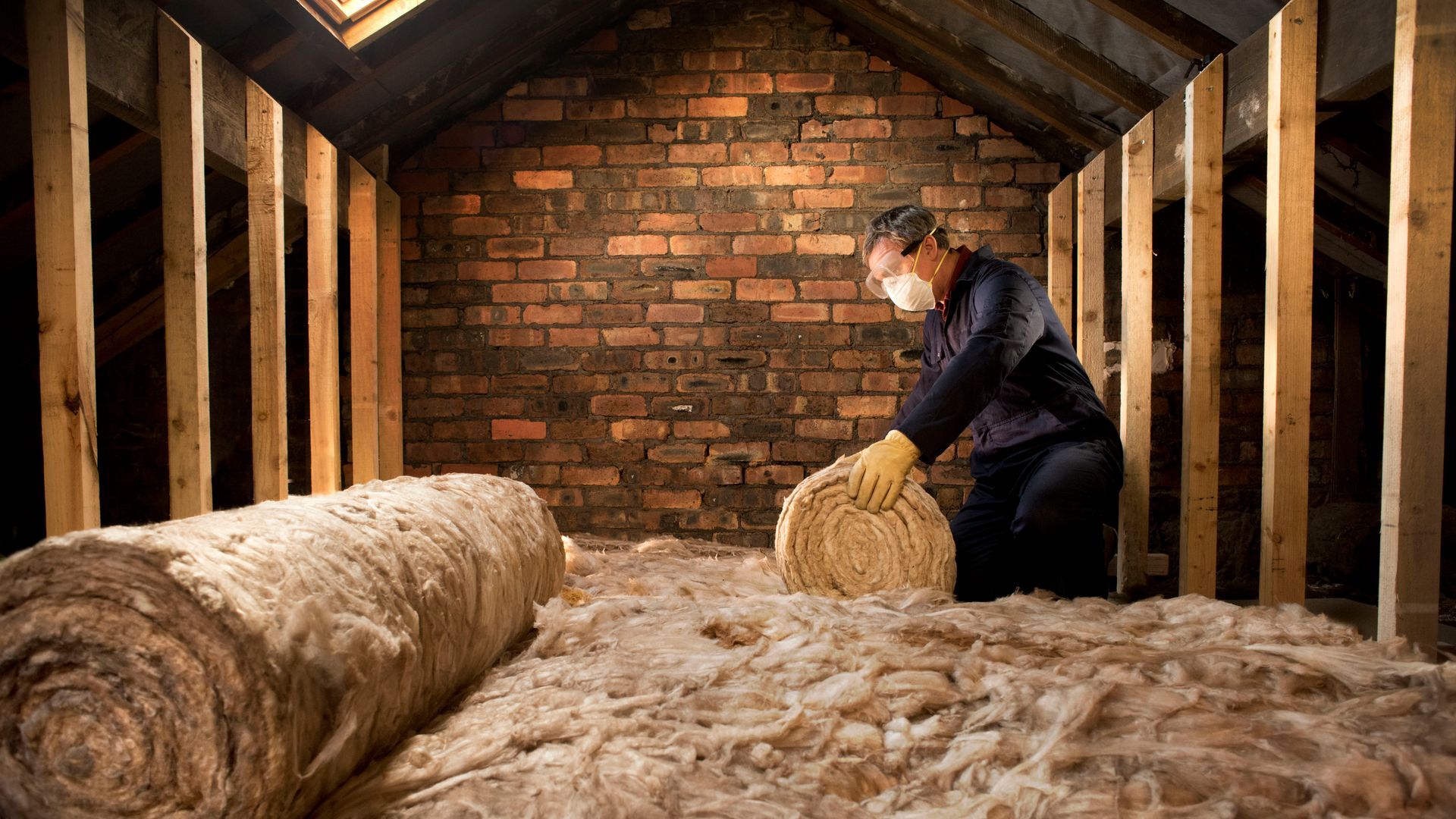 Person in attic installing insulation. Wooden beams, brick wall. Insulation is beige. Person is wearing a mask and gloves.