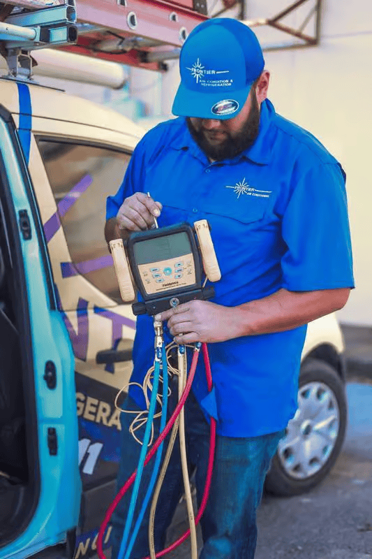 HVAC technician in blue uniform, examining equipment next to van. Red and blue hoses visible.