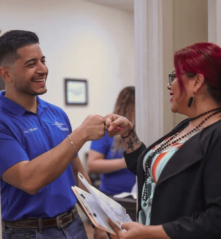 Man and woman bump fists, smiling. Man wears blue shirt, woman wears glasses and black blazer, holding paperwork.