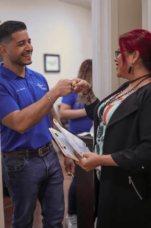 Man in blue shirt and woman in black blazer bump fists, possibly in an office setting.