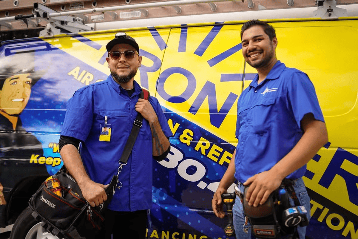 Two men in blue shirts pose in front of a colorful promotional banner.