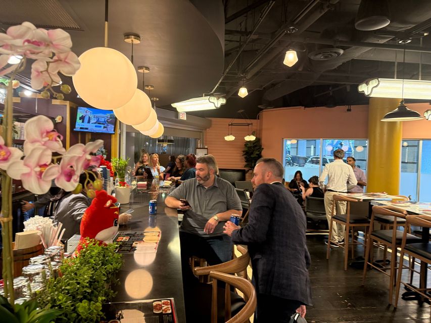 People chatting at a bar counter in a warmly lit restaurant with hanging pendant lights and floral decor