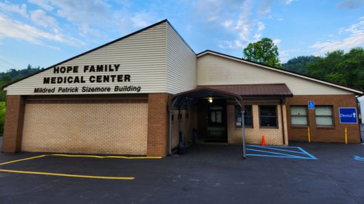 Brick building entrance with covered porch, parked car, and wooded hillside in the background