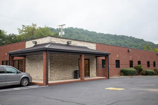 Brick building with covered entrance and parked car beside a wooded hillside