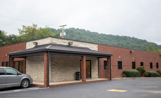 Brick building entrance with covered porch, parked car, and wooded hillside in the background