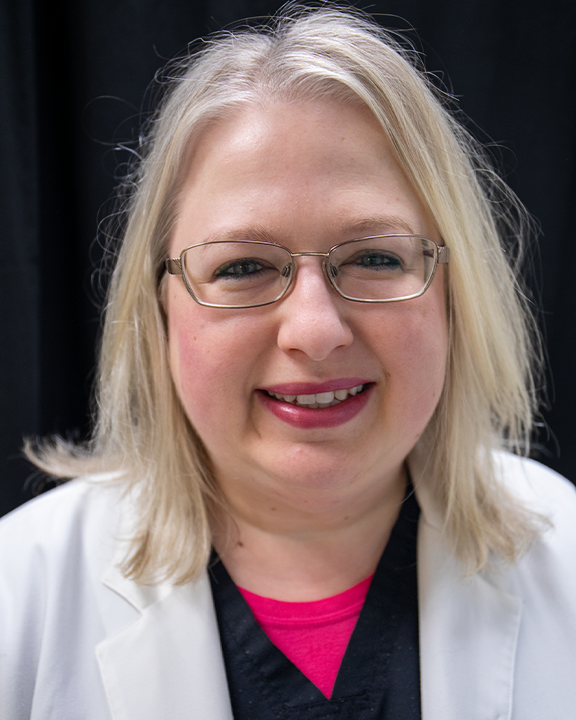 Portrait of a smiling person with blond hair and glasses, wearing a white coat over a pink top