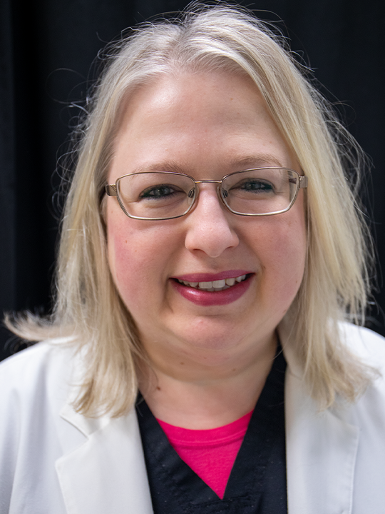 Smiling person with glasses and blonde hair in a white coat over a bright pink top